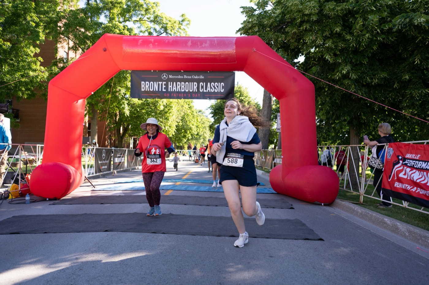 Two runners crossing the finish line under the red Bronte Harbour Classic arch at the Mercedes-Benz Oakville Bronte Harbour Classic 5K, Bronte Heritage Waterfront Park, Oakville